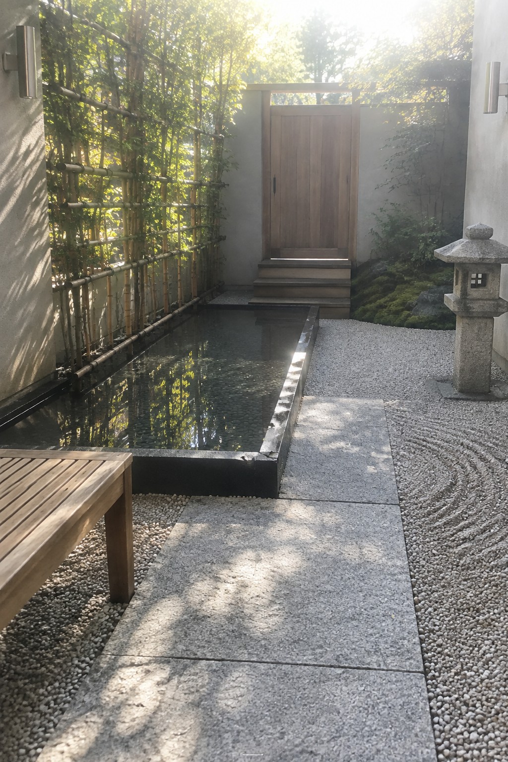 Large rectangular stone slabs form a straight path through a gravel courtyard beside a narrow reflecting pool and bamboo fence.