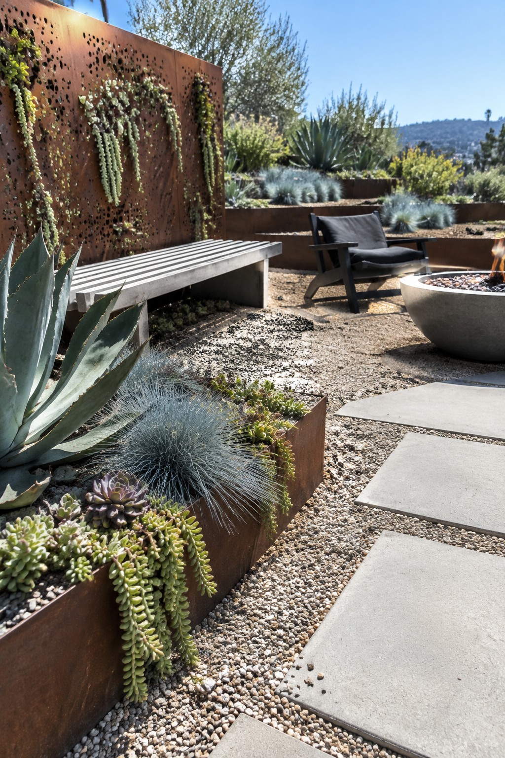 A gravel patio has rust-colored raised planters filled with succulents, a concrete path, a bench, a chair, and a round fire pit.