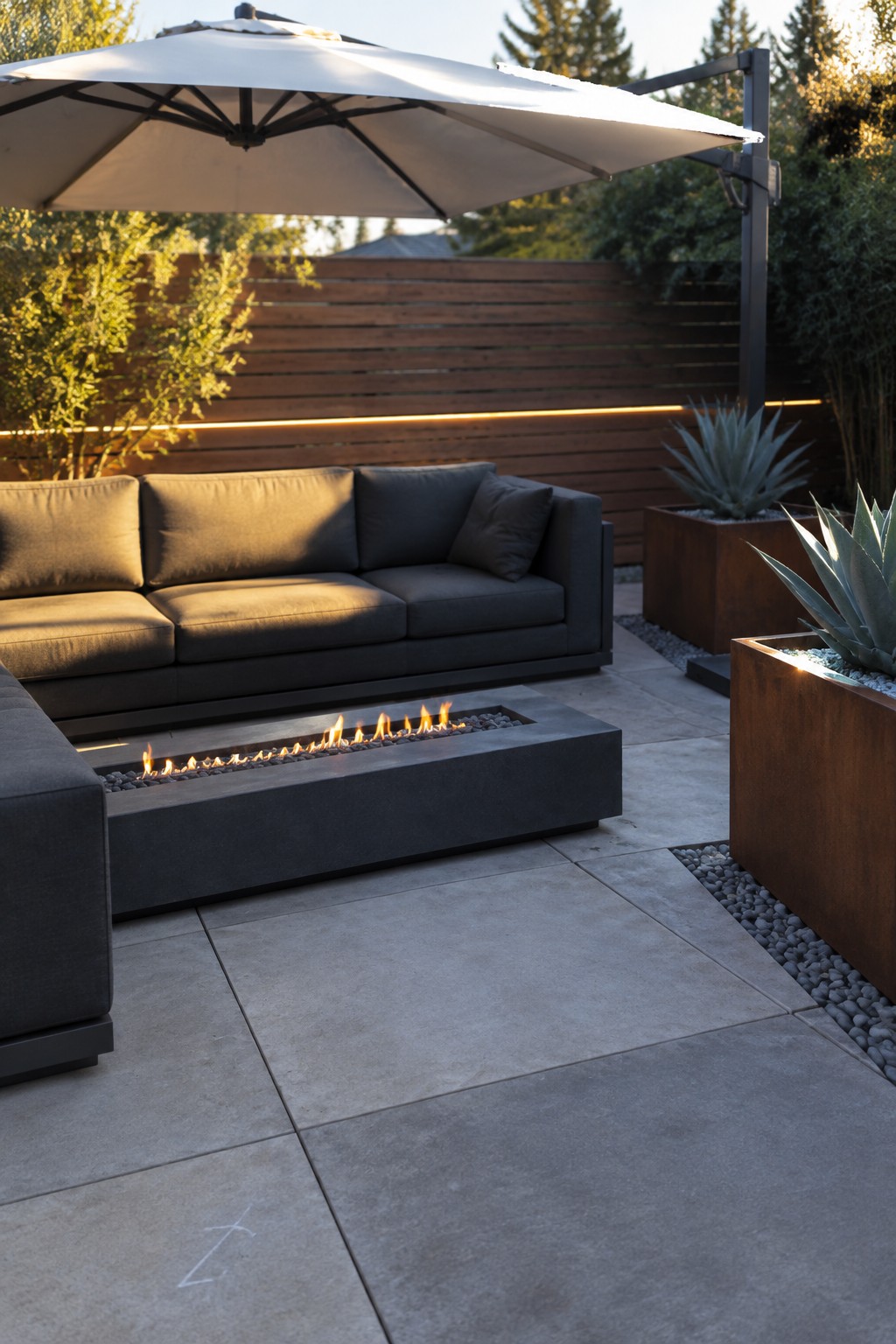 A rectangular gas fire pit sits in front of a dark outdoor sofa on a paved patio beneath a large umbrella.