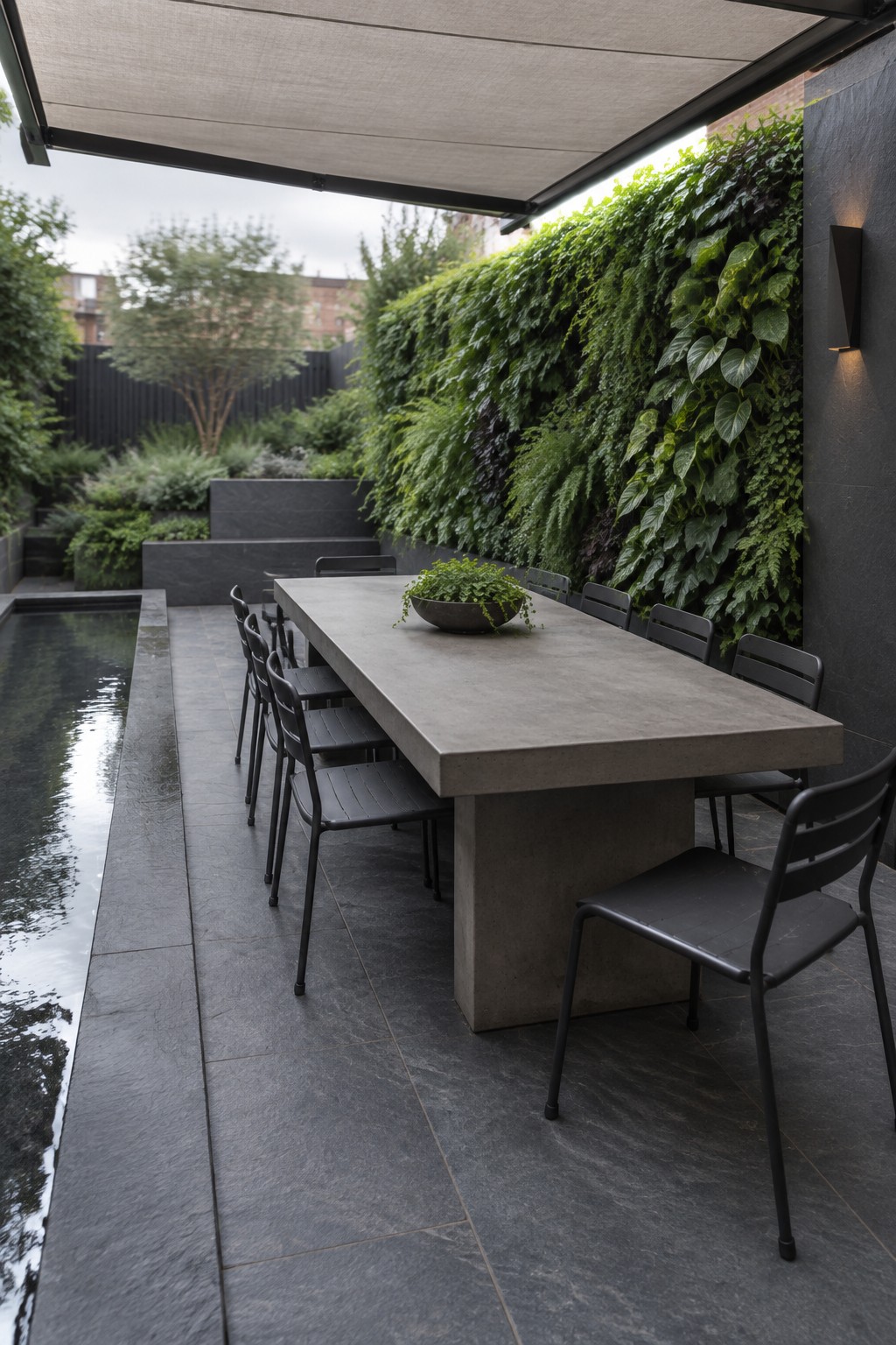A long concrete dining table with black chairs sits on a tiled patio beside a narrow pool and a leafy vertical garden.