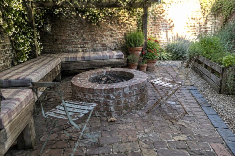 20 Rustic Brick Patio Ideas on a Budget Using Reclaimed Materials A circular brick fire pit sits in a brick-paved courtyard with cushioned bench seating and metal chairs.