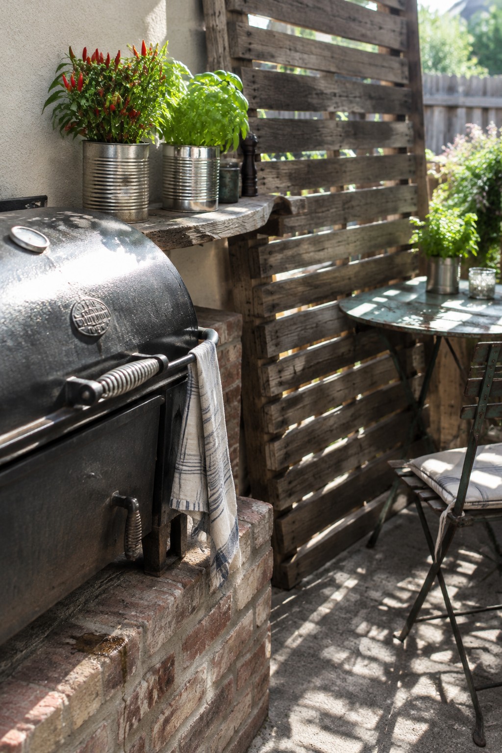 A rustic backyard patio with a weathered wooden privacy screen, a brick grill base, potted plants, and a small metal table and chairs.