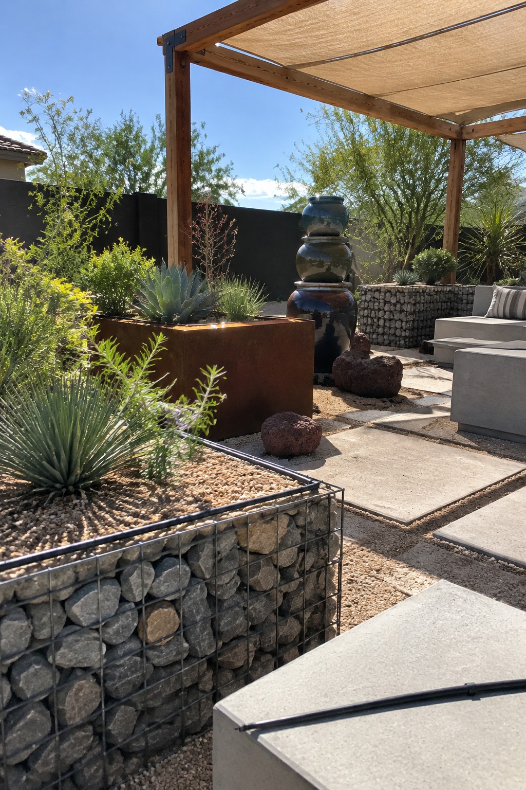 A gravel-filled gabion raised bed planted with succulents sits beside concrete patio pavers under a wooden pergola.
