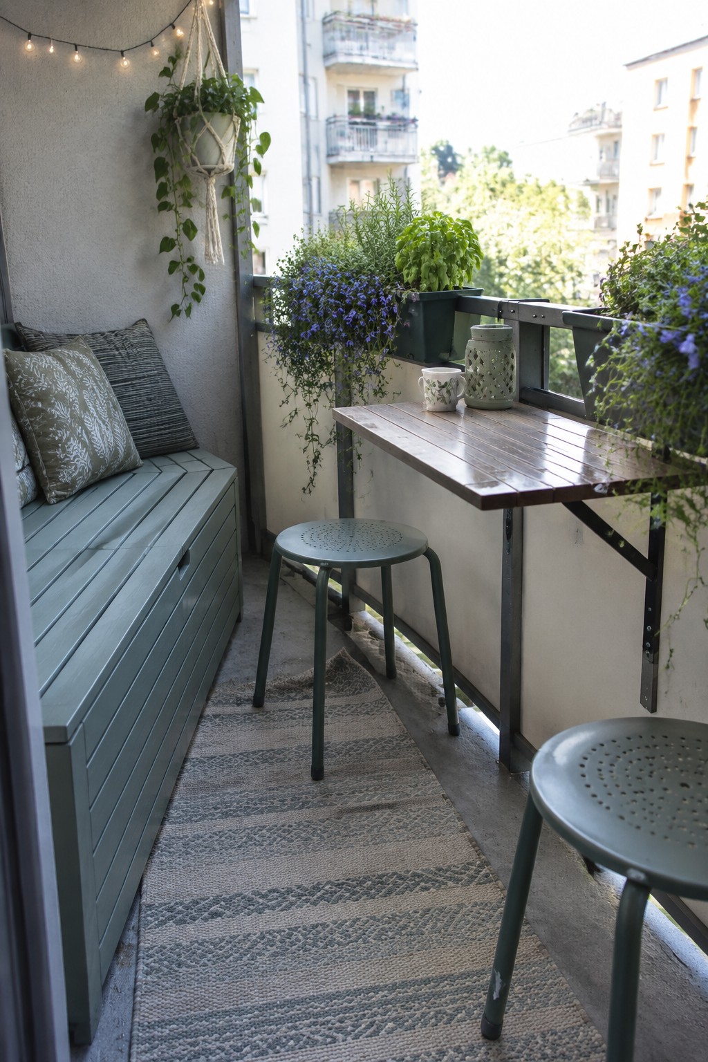 A narrow balcony with a wooden bar-height table, metal stools, a built-in bench, and potted plants.