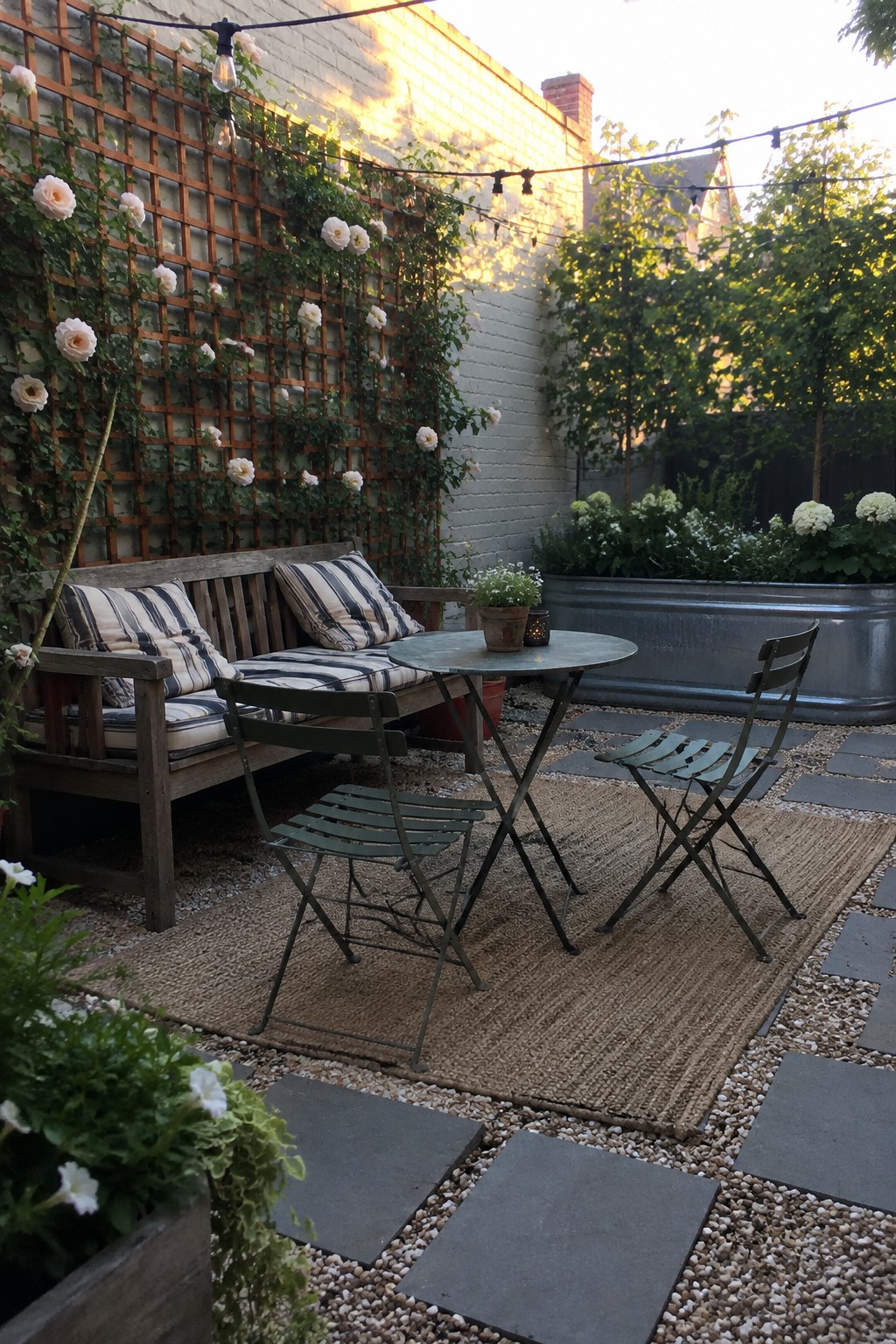 A wooden trellis covered with pale roses stands behind a cushioned bench and small patio table.