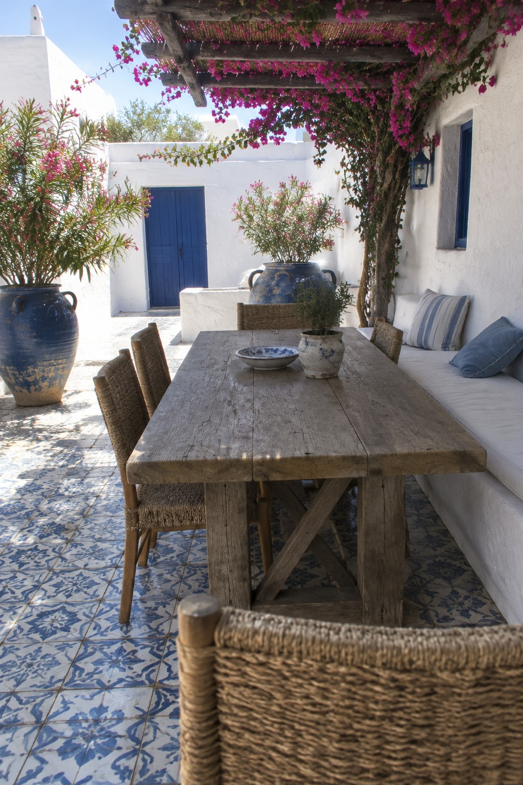A long wooden dining table and woven chairs sit on a blue-and-white tiled patio beside a built-in bench, beneath a pergola covered in pink flowers.