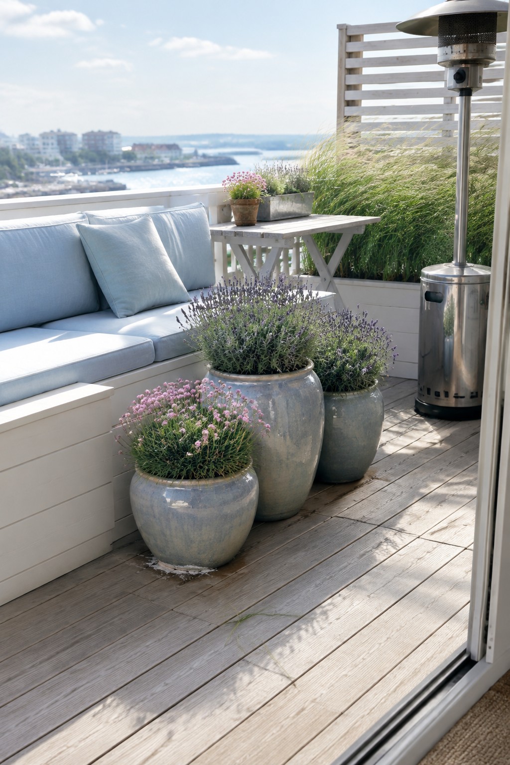 Three large ceramic pots of flowering plants sit beside a cushioned outdoor sofa on a wooden balcony.