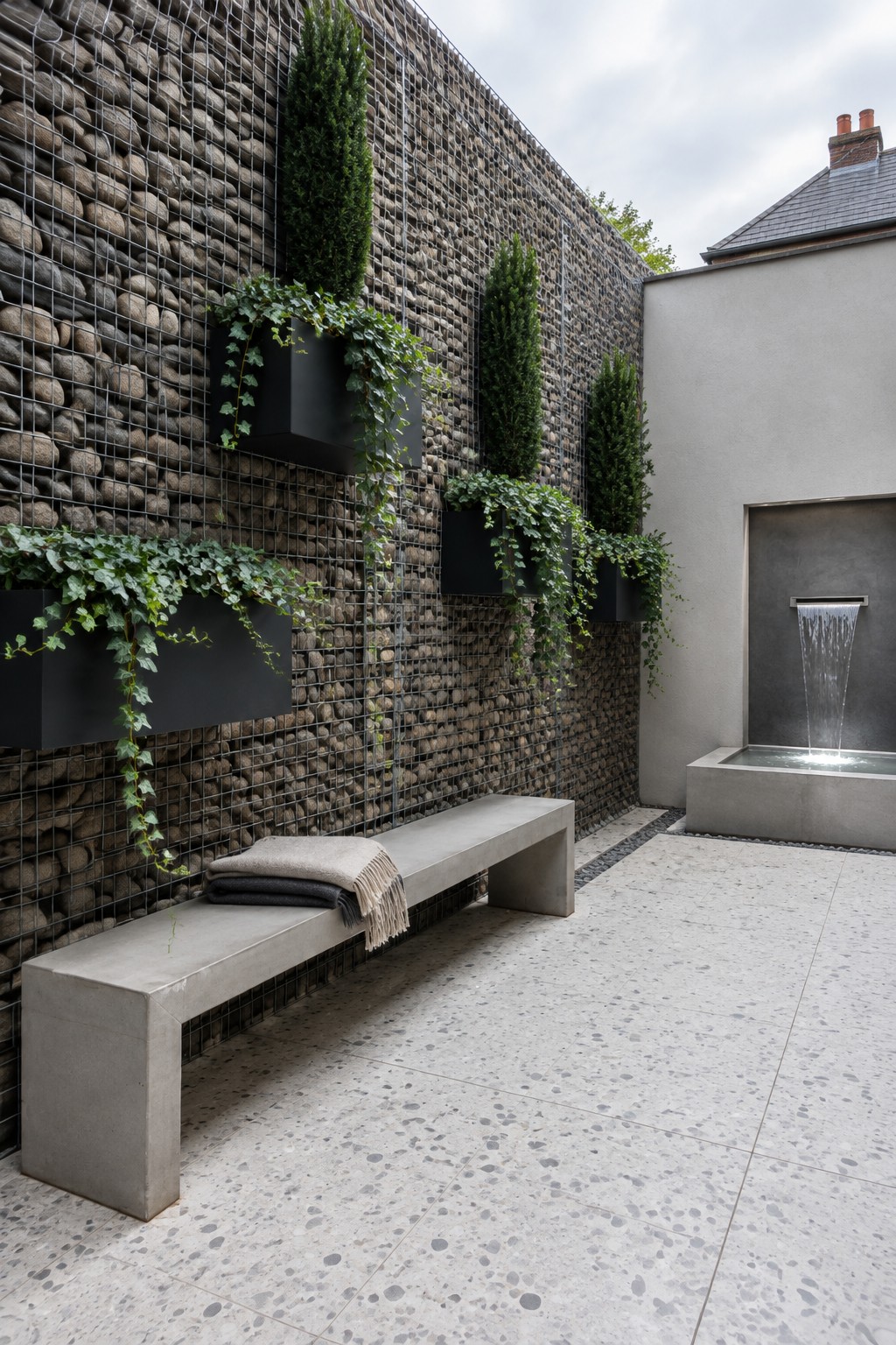 A stone-filled gabion wall with mounted planters stands beside a concrete patio bench and a small wall fountain.