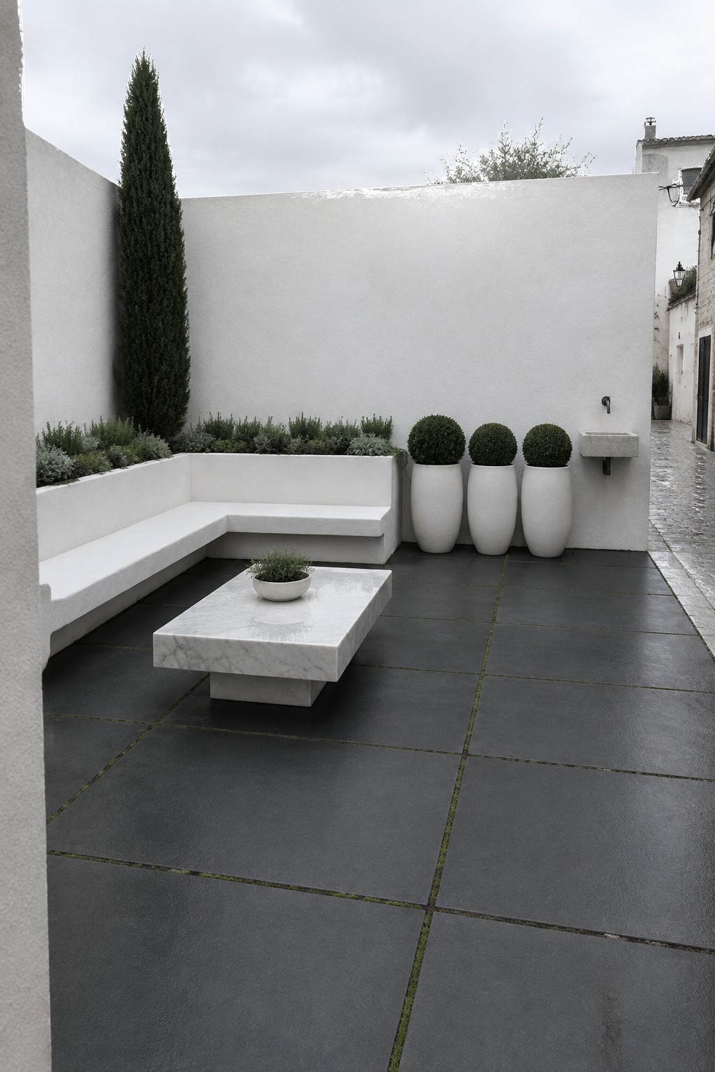 A white L-shaped built-in bench sits beside dark square patio tiles, with a low marble table and three potted shrubs.